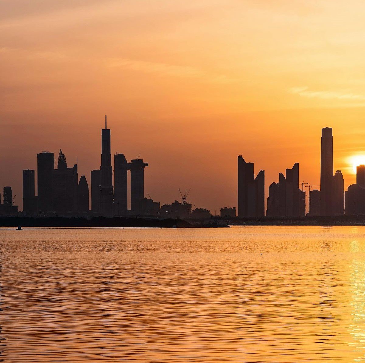 Modern Abu Dhabi skyline silhouetted against a vivid golden sunset, reflected across calm waterfront waters.