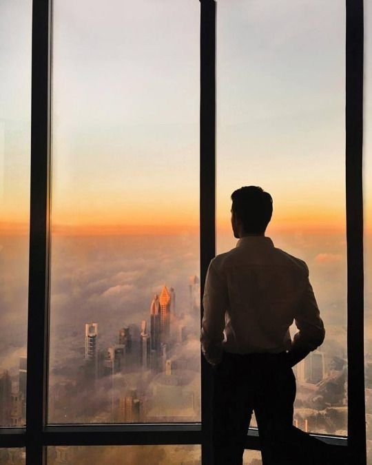 Silhouette of a businessman standing beside floor-to-ceiling windows at sunset, overlooking a modern city skyline rising through clouds.