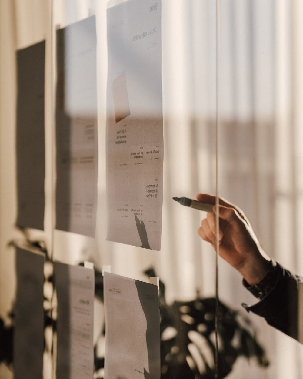 Hand using a marker to review printed planning documents displayed on a glass wall in a modern workspace.