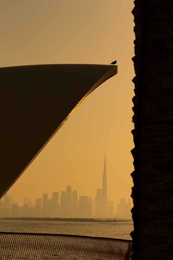 Dubai skyline and Burj Khalifa seen across the water at golden hour, framed by dramatic contemporary architecture and a palm trunk.